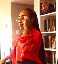 African-American woman in red blouse with glasses stands smiling in front of a bookcase. 