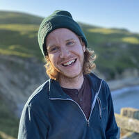 White mail with blonde, curly hair wearing a green beanie and a blue pullover standing near a cliff overlooking the ocean.