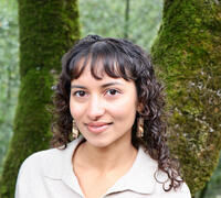 Asian female with shoulder length curly brown hair and bangs standing in front to two moss covered trees. 