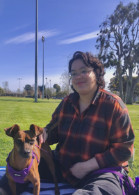 Photo of a young woman wearing jeans and a red and black checkered shirt sitting in the grass with a small brown dog