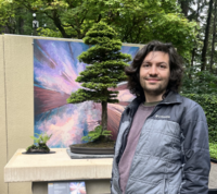 Man with long brown hair wearing a dark jacket standing in front of a piece of art with a miniature tree. 