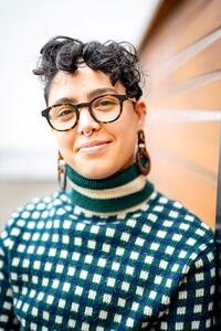 Young person with short, dark, curly hair wearing glasses and a turtleneck sweater and large earrings.