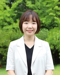 Asian woman with short dark hair wearing a white blazer and black shirt standing outside near some trees. 